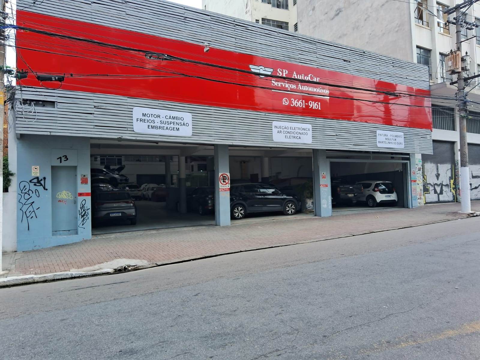 Fachada da oficina no número 73, com três boxes abertos, veículos dentro e placas de motor, câmbio, freios, suspensão e embreagem.